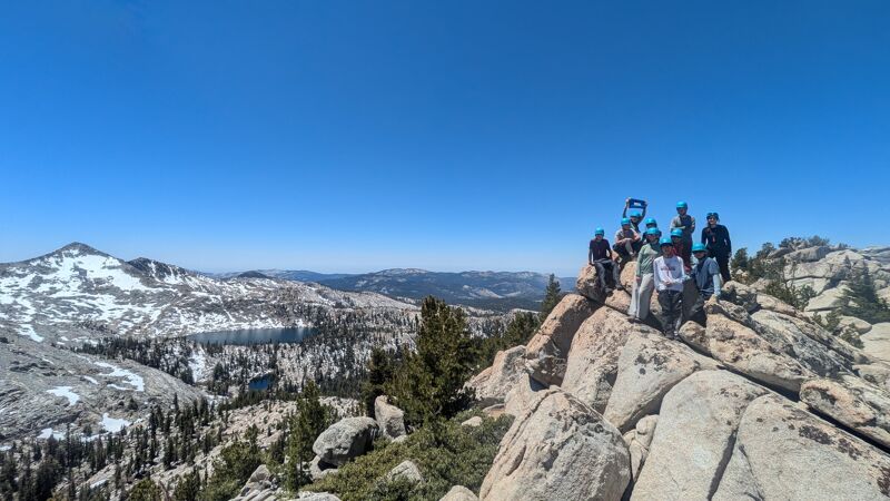 A group of people stands atop a rocky mountain peak on a clear, sunny day. The landscape features snow-capped mountains in the distance, interspersed with patches of trees and a body of water. The foreground is dominated by large, light-colored boulders and sparse vegetation, contrasting with the vast, open sky above.